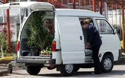 White Piaggio Porter van with rear doors open, displaying potted plants inside, parked in front of red industrial buildings.
