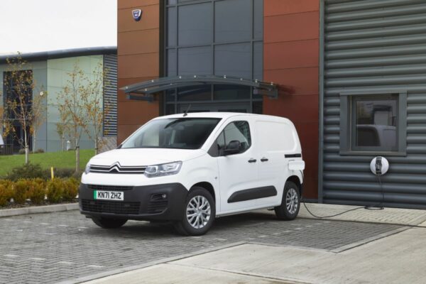 White Citroën ë-Berlingo electric van parked outside modern building, charging cable connected to wall-mounted charger.