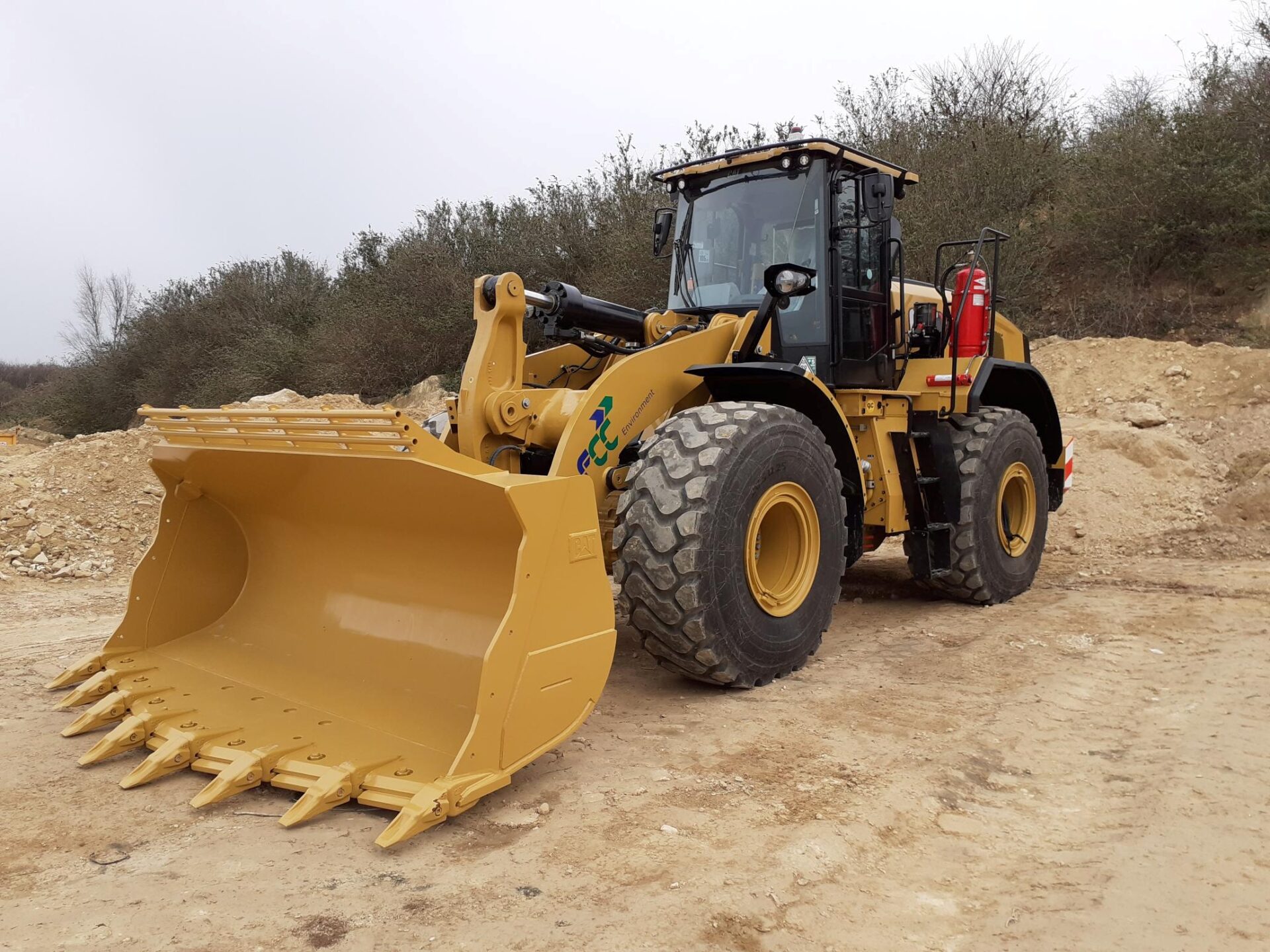 Yellow wheel loader with bucket attachment on gravel site surrounded by bare trees.