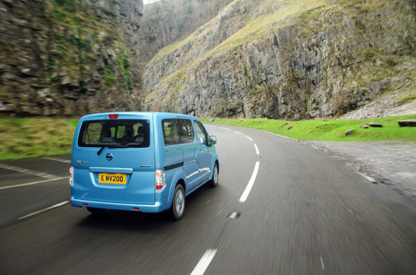 Blue Nissan e-NV200 Evalia driving on mountain pass road with steep rocky cliffs either side.