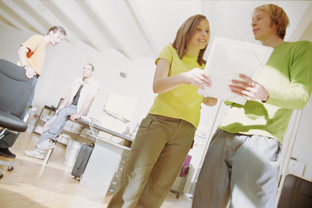 Woman in yellow top discusses financing options with man in green jumper whilst colleagues work in bright office space.