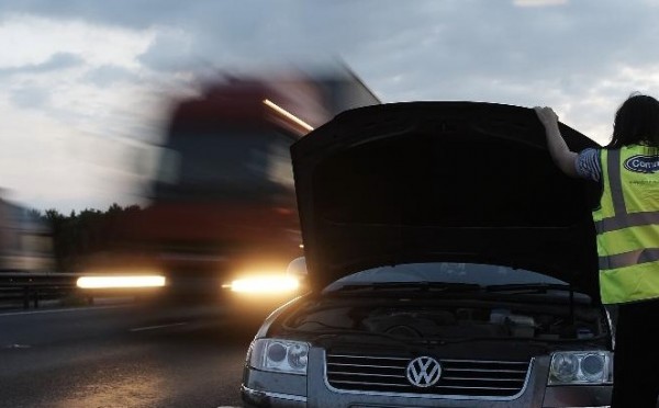 Broken-down Volkswagen van on motorway with HGV truck approaching and recovery worker in high-visibility jacket nearby.