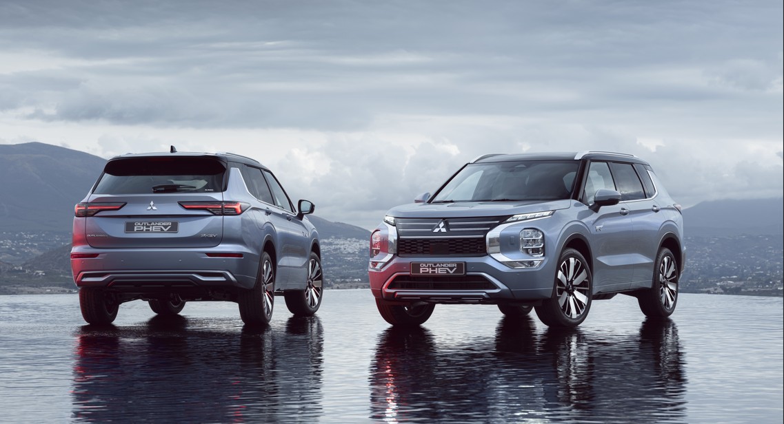 Two Mitsubishi vehicles parked on wet surface with mountains and cloudy sky behind them: silver Outlander PHEV shown from…