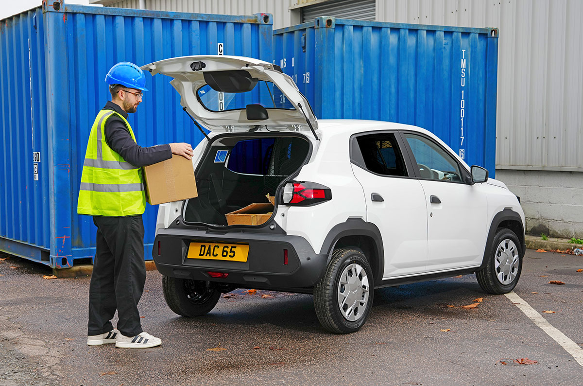 Worker in high-visibility clothing loading cardboard box into white Dacia Spring Cargo electric van with tailgate open,…