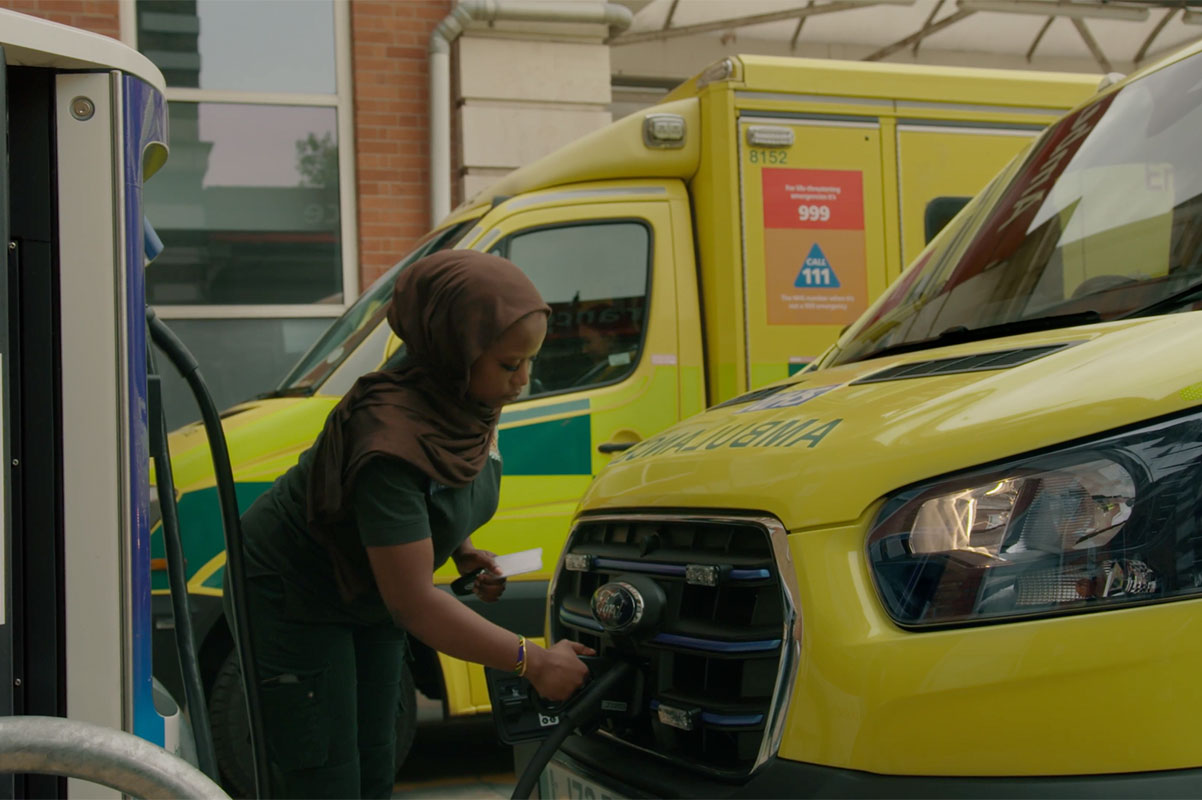 Paramedic in green uniform sitting in driver's seat of yellow electric ambulance, charging vehicle at station.