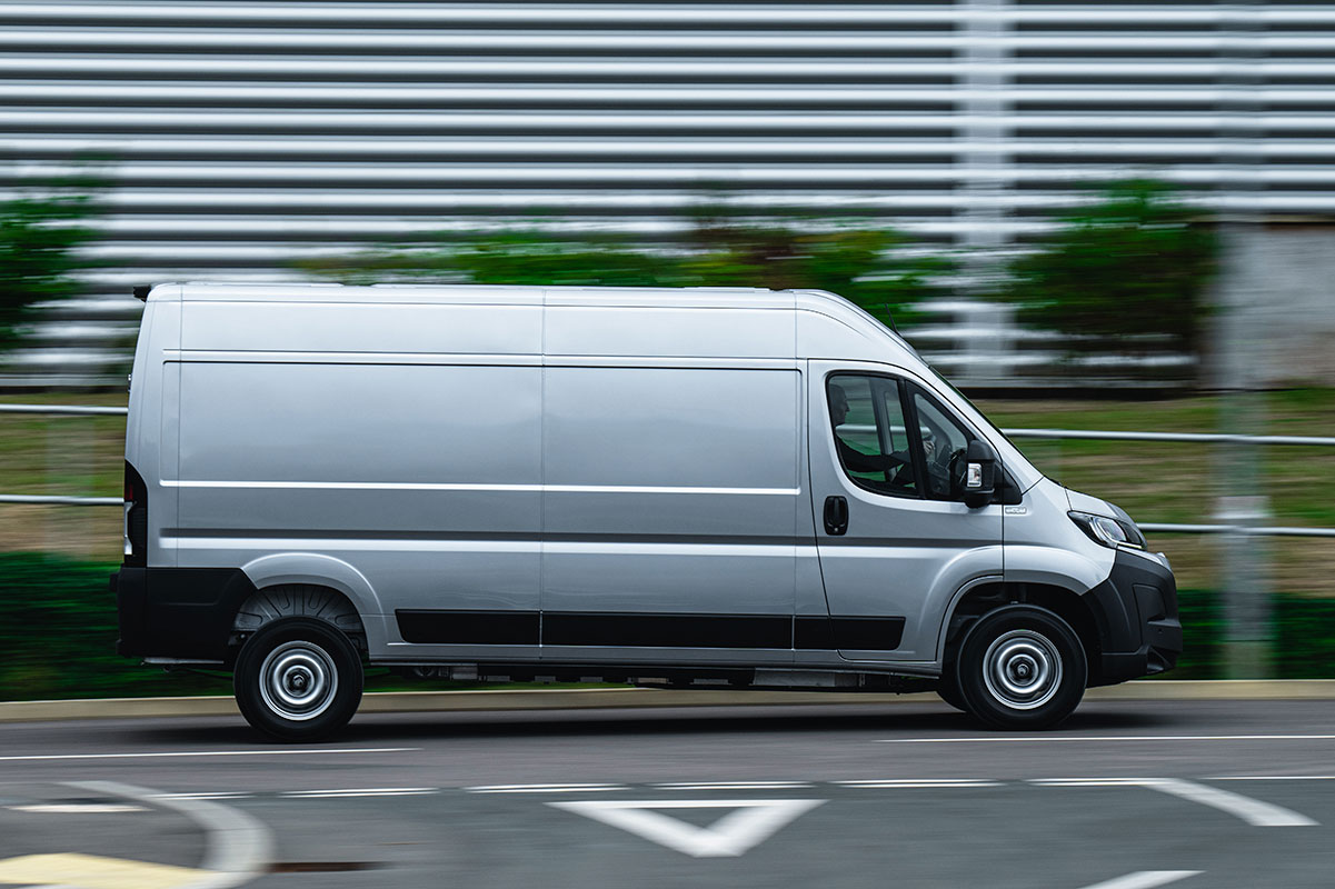 White commercial van driving on a road with blurred background and road markings visible.