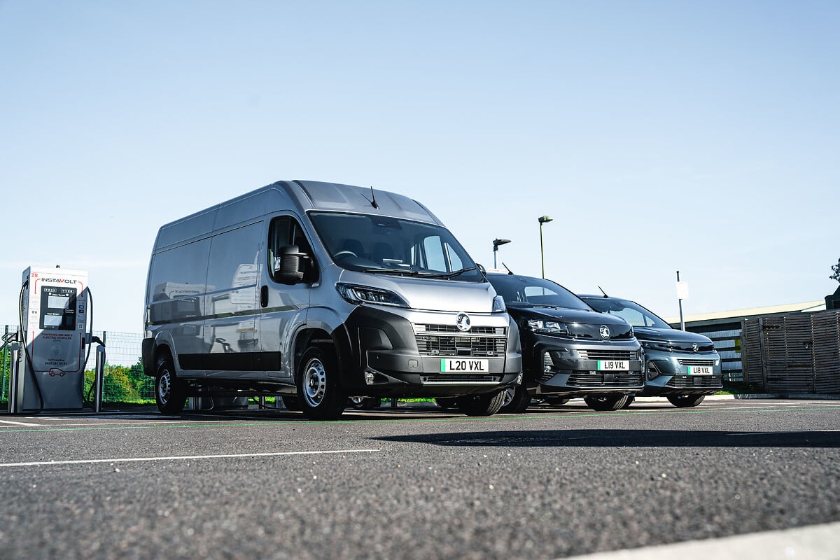 Vauxhall electric van parked alongside diesel vans at a charging point in an industrial yard.