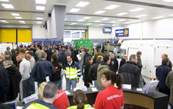 Crowded indoor trade show or exhibition hall with numerous visitors browsing at display stands and booths.