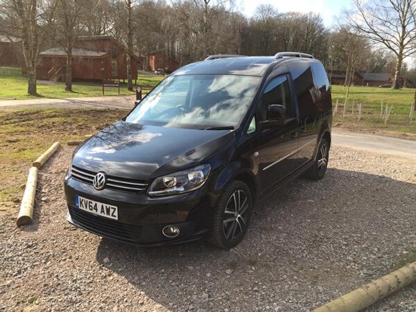 Black Volkswagen Caddy van parked on gravel drive with bare trees and farm buildings in background.