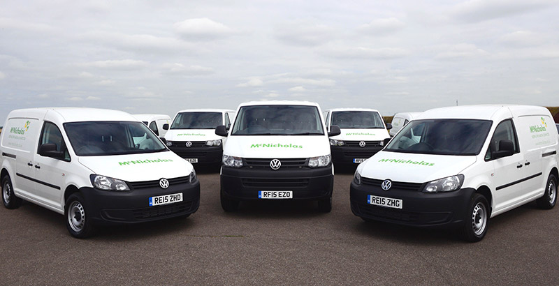Fleet of five white Volkswagen vans with green McNicholas branding parked in a line, with central Volkswagen Transporter van.