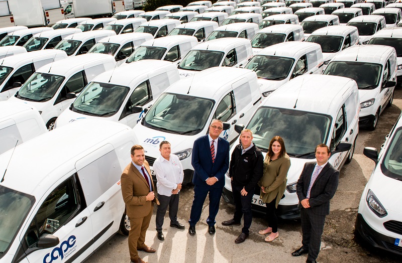 Six business people stand among rows of white commercial vans in a fleet yard, discussing the cost savings from switching…