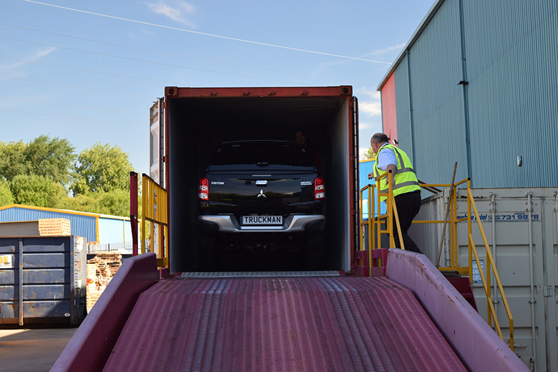 Dark Mitsubishi L200 pickup loaded into burgundy Truckman hardtop truck bed at industrial yard with worker in…