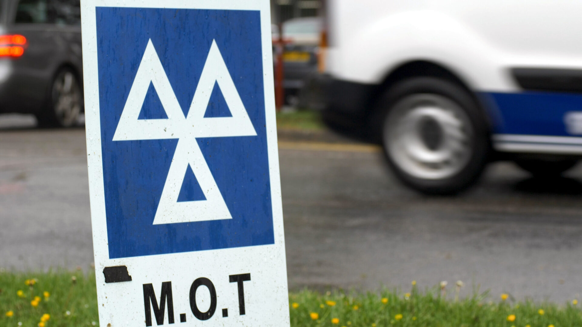 MOT sign with white triangular logo on blue background, parked cars in background