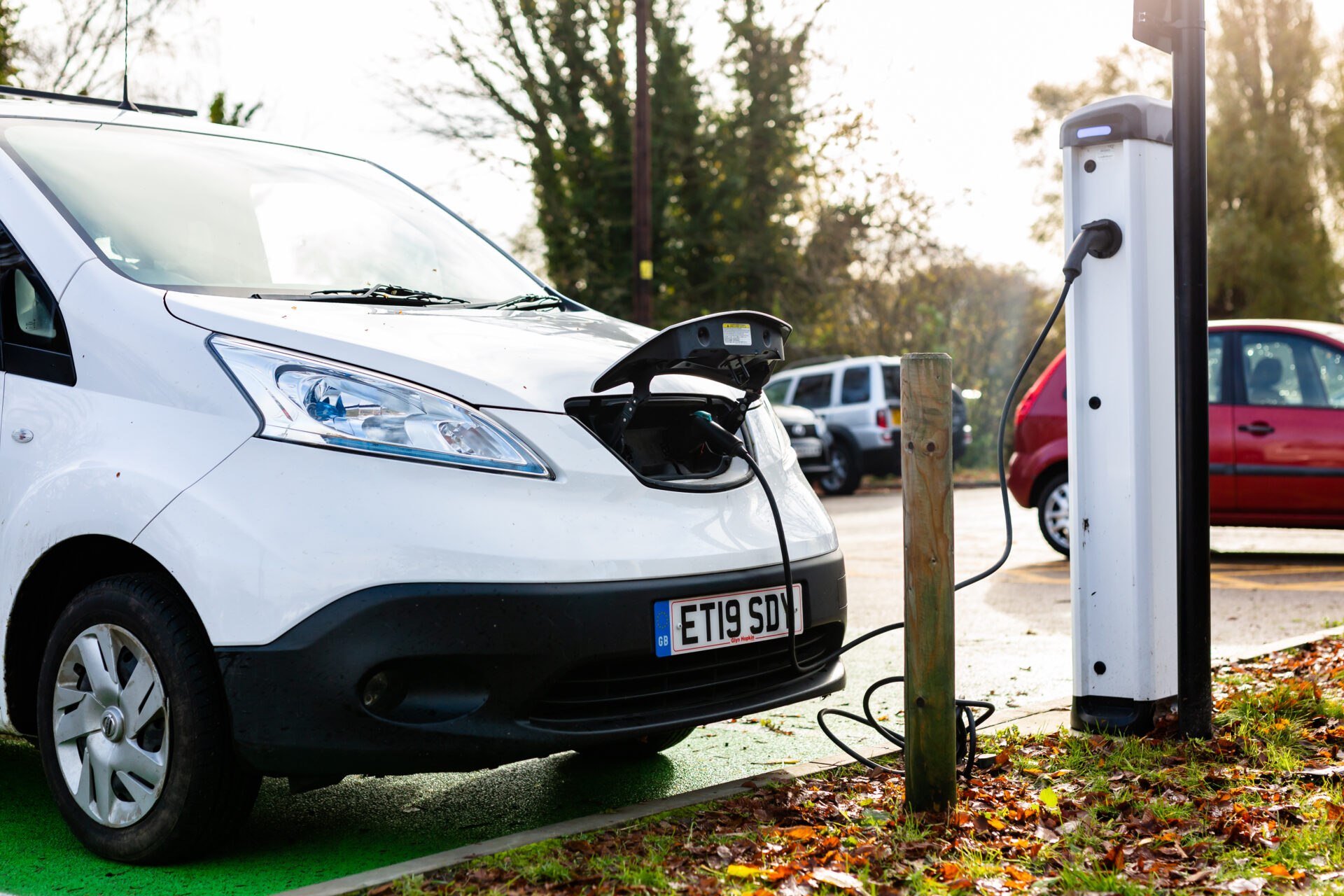 White electric van charging at public charging point in car park with other vehicles in background.