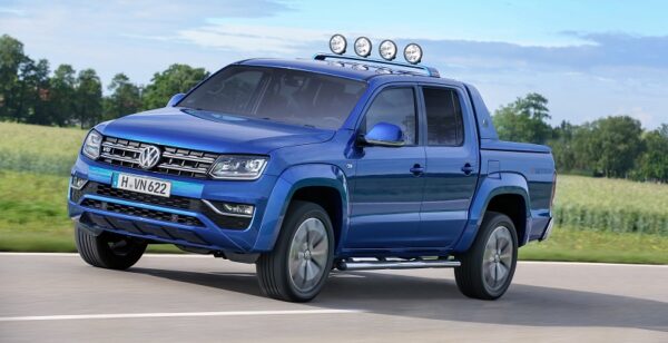 Blue Volkswagen Amarok double cab pick-up truck with roof-mounted spotlights driving on a road with green fields in…