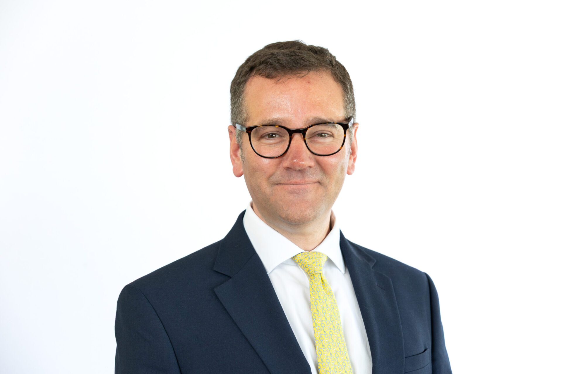 Man wearing dark suit, white shirt, and yellow tie with glasses against white background, professional headshot.