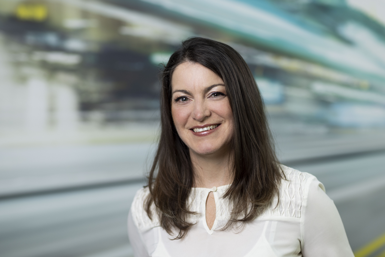 Woman with long dark hair wearing white blouse, smiling at camera in front of blurred industrial background.
