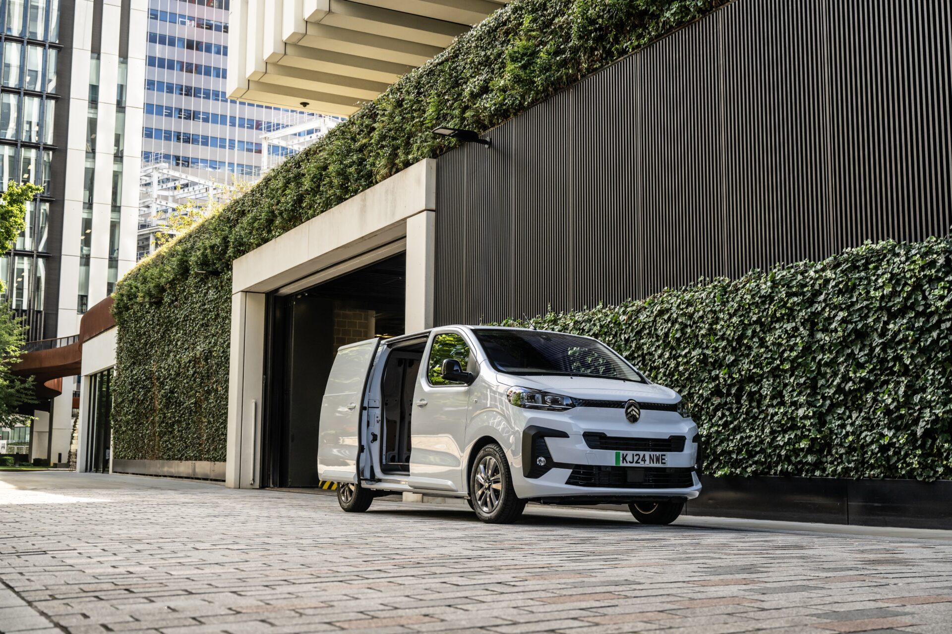 White electric van with open side door parked outside modern building with ivy-covered walls and garage entrance.