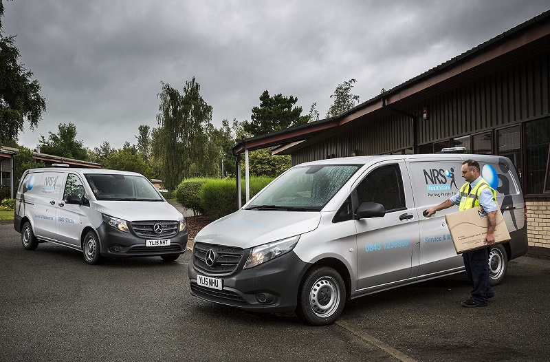 Two white Mercedes-Benz vans parked outside a brick building, with NHS worker unloading boxes from the nearest vehicle.