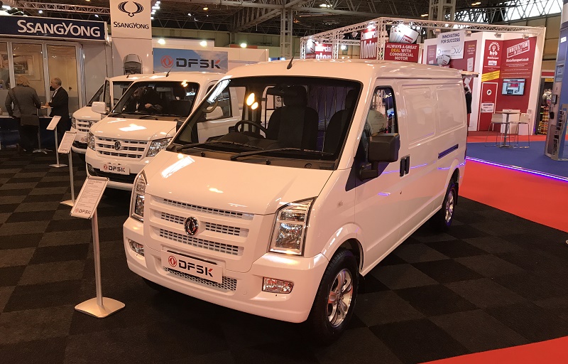 White DFSK commercial van displayed at trade show with open side door, other vehicles and exhibition stands visible behind.