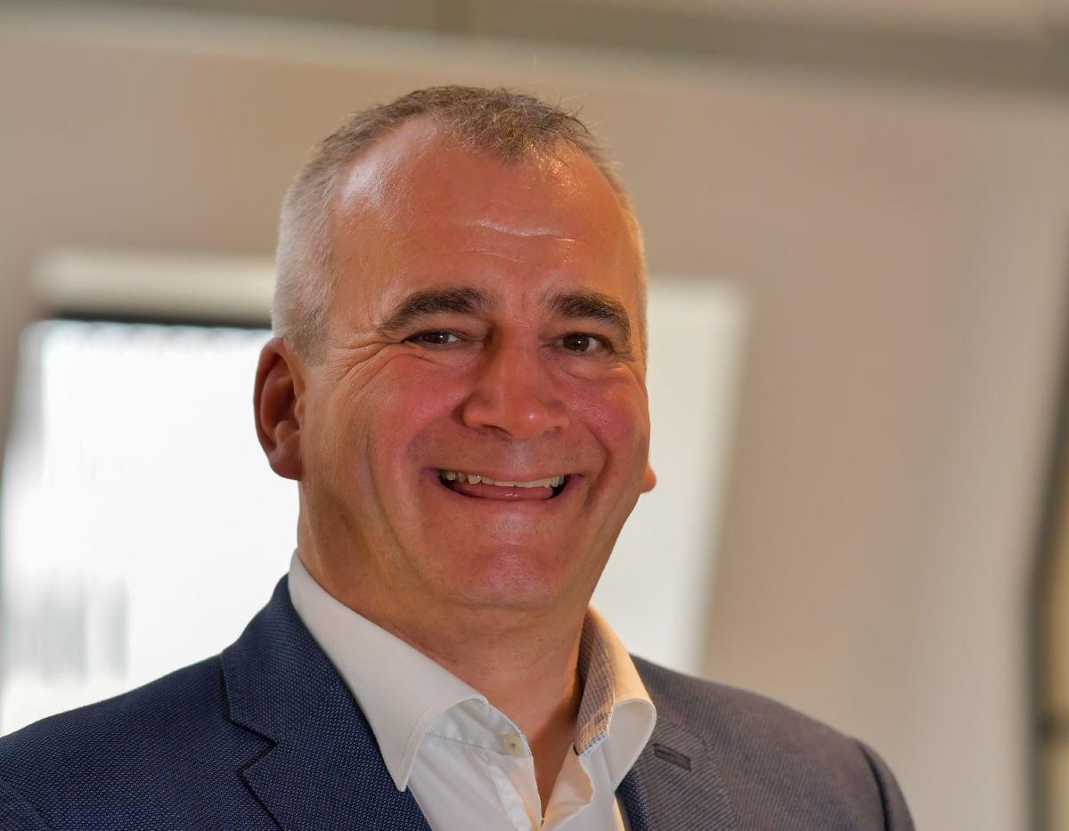 Middle-aged man with grey hair smiling at camera, wearing navy blazer and white shirt in office setting.