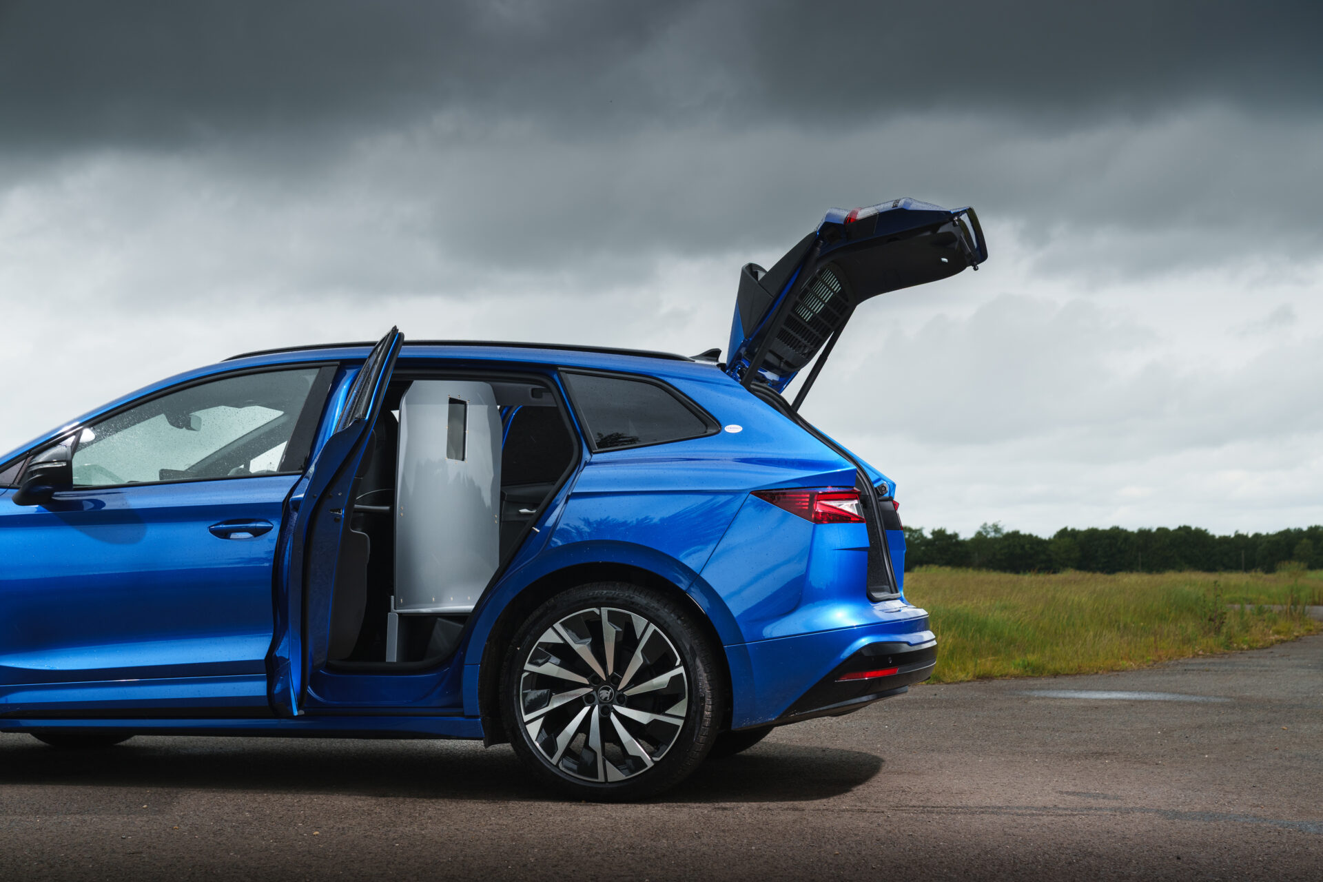 Blue Škoda Enyaq Cargo electric vehicle with open side door and elevated roof hatch, shown in profile on tarmac with…