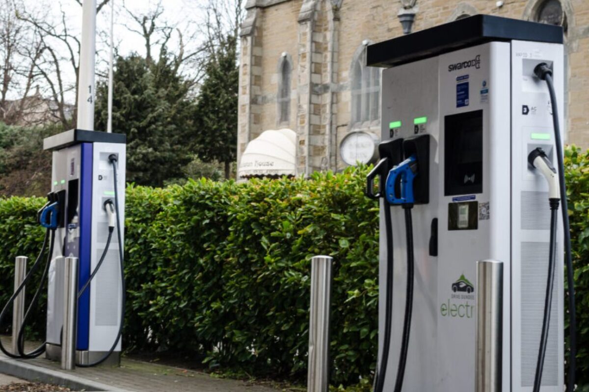 Electric vehicle charging points in a public car park beside a church and hedge, with stone bollards and green indicator…