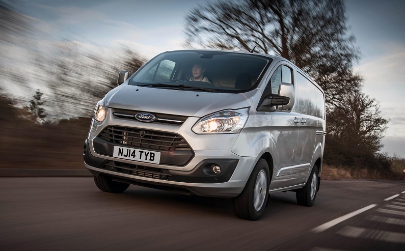 White Ford Transit Custom van driving on road with bare trees in background