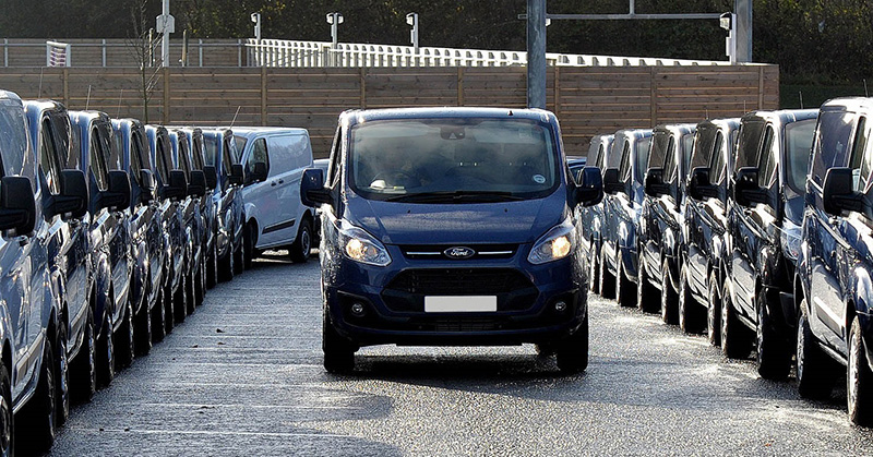 Blue Ford van driving down centre aisle of large vehicle storage lot lined with white vans on both sides.