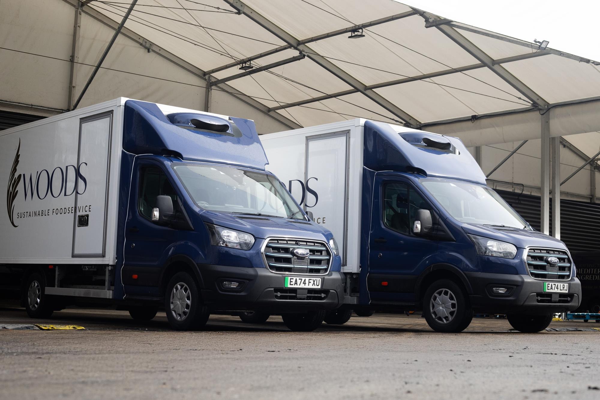 Two blue and white Ford E-Transit vans parked under a canopy, displaying Woods Foodservice branding.
