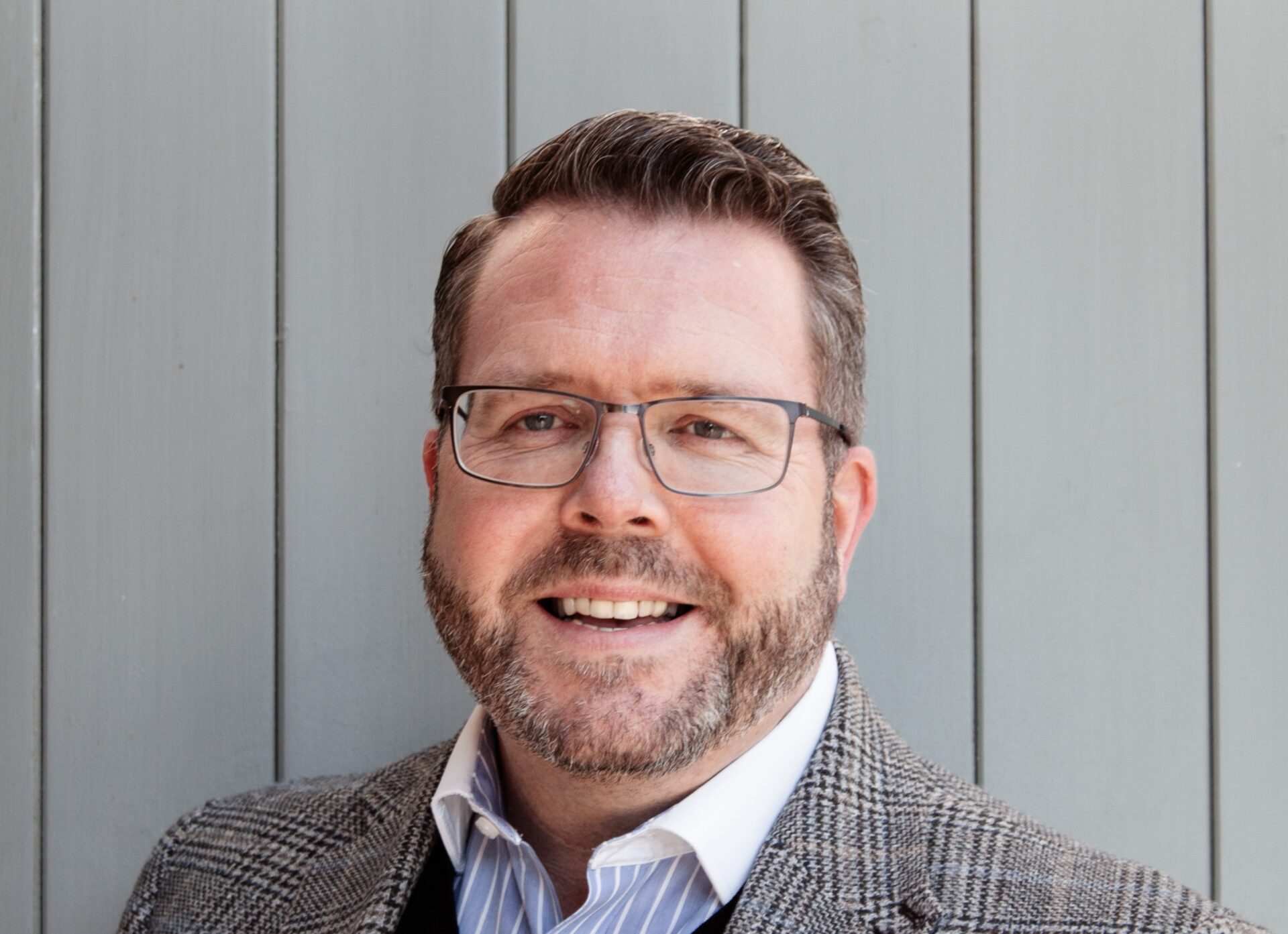 Man with glasses and beard wearing grey blazer and blue striped shirt, smiling at camera against grey corrugated backdrop.