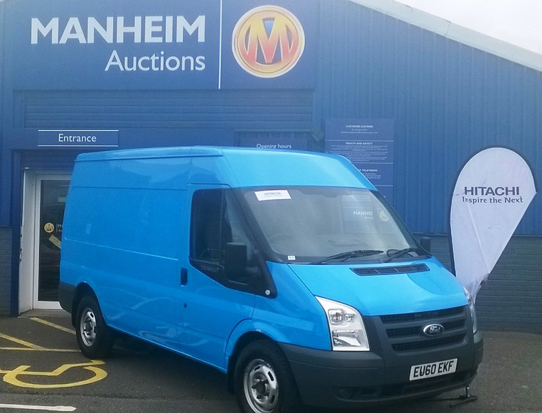 Blue Ford Transit van parked outside Manheim Auctions building with Hitachi branding visible.