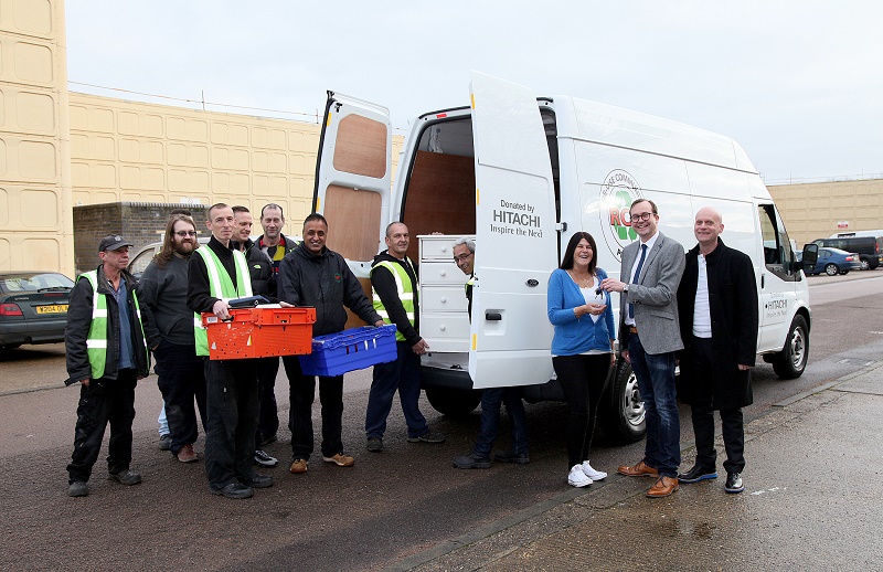 Group of staff and volunteers standing beside white Hitachi Transit van, holding red and blue storage boxes.