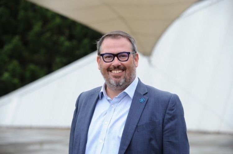 Man in blue blazer and glasses smiling at camera with modern building in background.