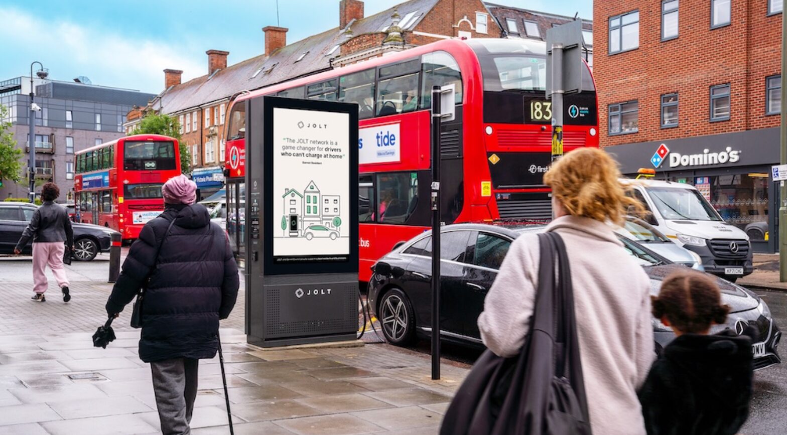 Red JOLT ad-funded electric vehicle charging station in busy urban high street with pedestrians and red bus visible in…