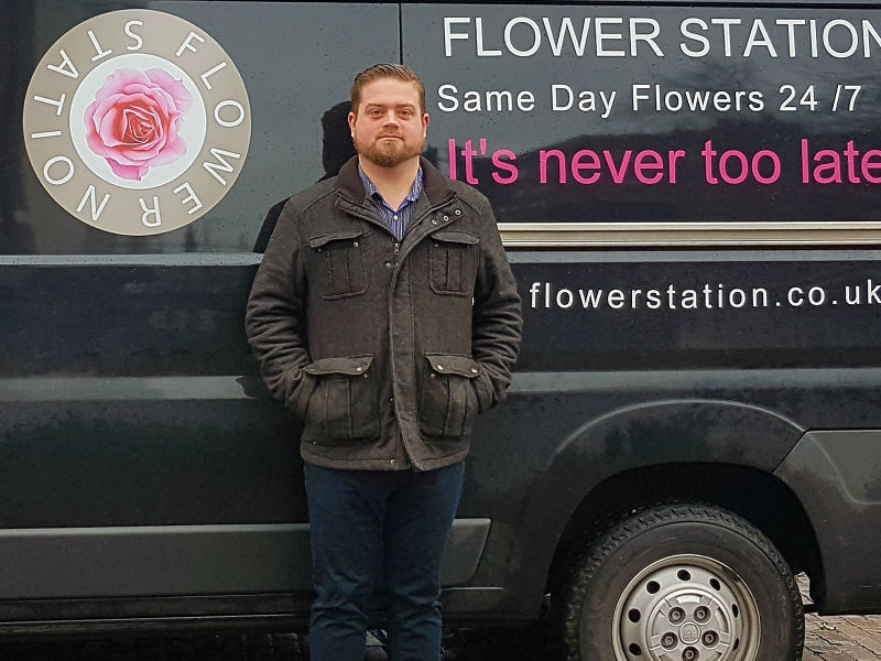 Man in dark jacket standing in front of Flower Station delivery van with pink rose logo and "Same Day Flowers 24/7" text.