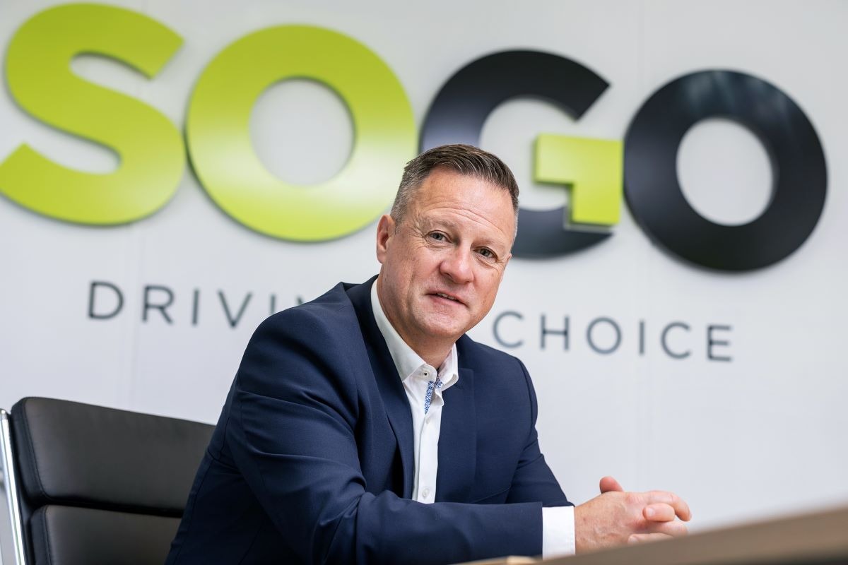 Man in dark suit seated at table with SOGO Driving Choice logo behind him, gesturing whilst speaking.
