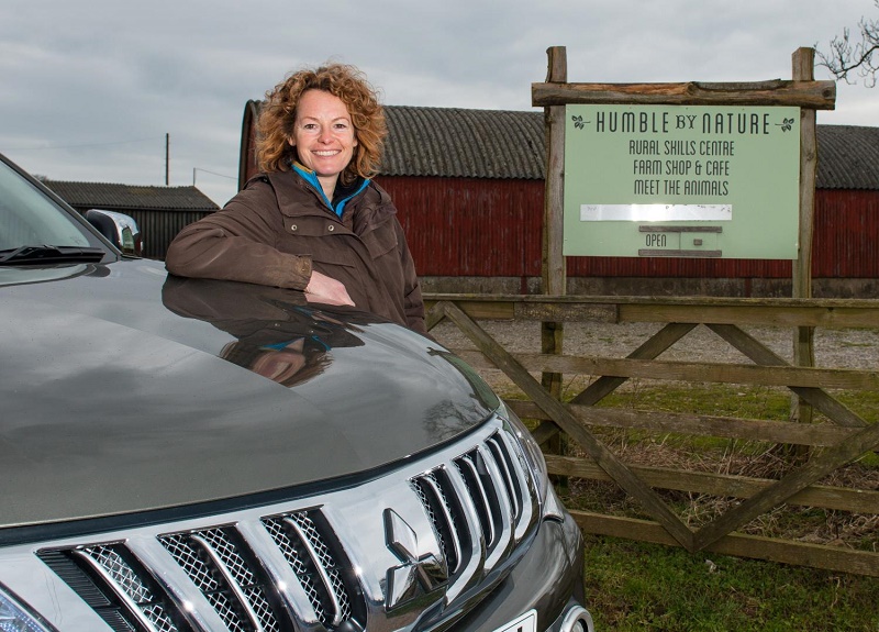 Woman with curly auburn hair leaning against a silver Mitsubishi L200 pickup truck beside a wooden gate and "Humble by…