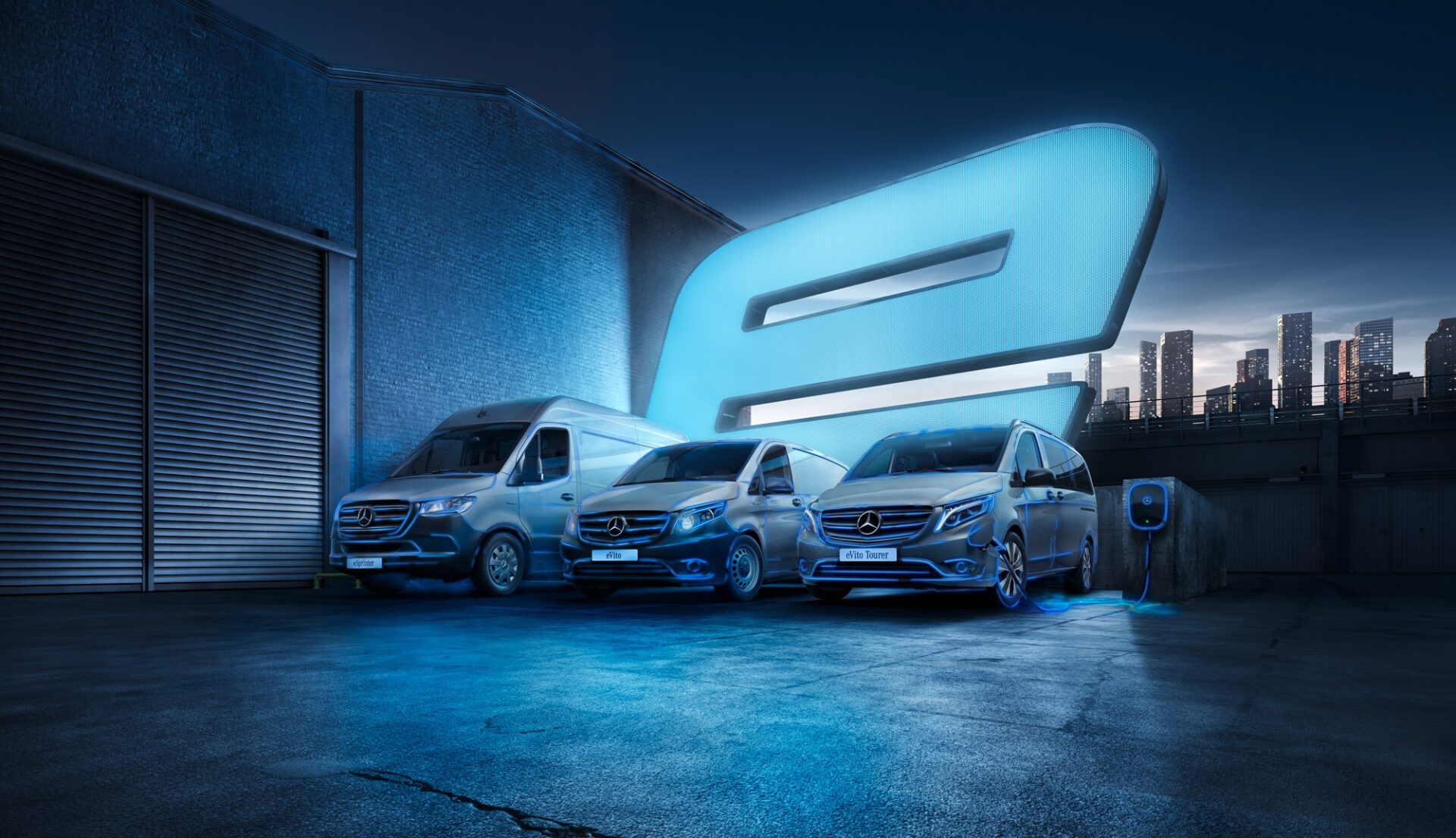 Three Mercedes-Benz vans parked in industrial setting with large glowing "e" symbol and charging point, city skyline behind.