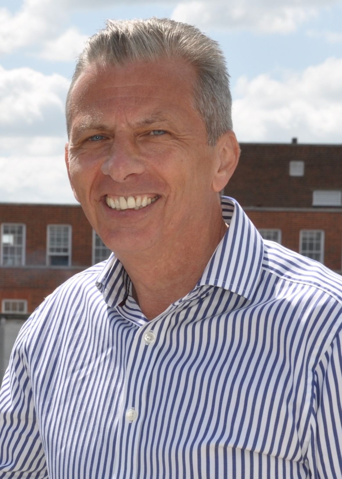 Middle-aged man with grey hair wearing blue and white striped shirt, smiling at camera with brick building behind.