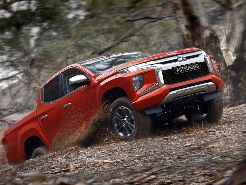 Red Mitsubishi L200 pickup truck climbing steep rocky terrain in woodland setting.