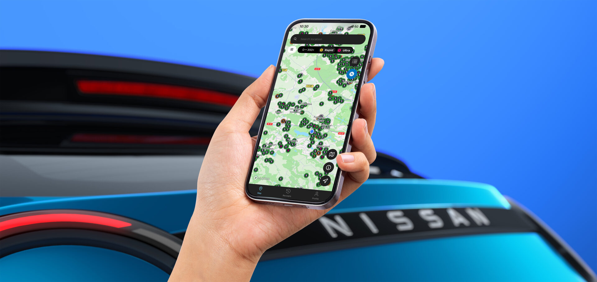 Hand holding smartphone displaying green charging location map, with Nissan electric vehicle in background against blue sky.