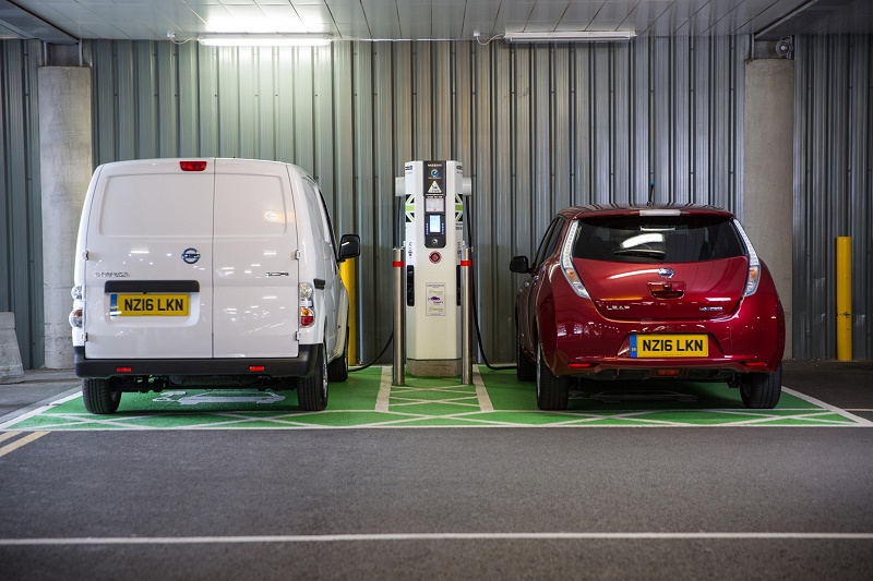White electric van and red electric car charging at shared fast-charging station in garage, demonstrating EV charging…