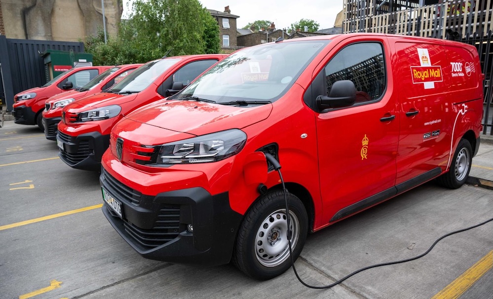 Line of red Royal Mail electric vans charging at a depot, with urban buildings in background.