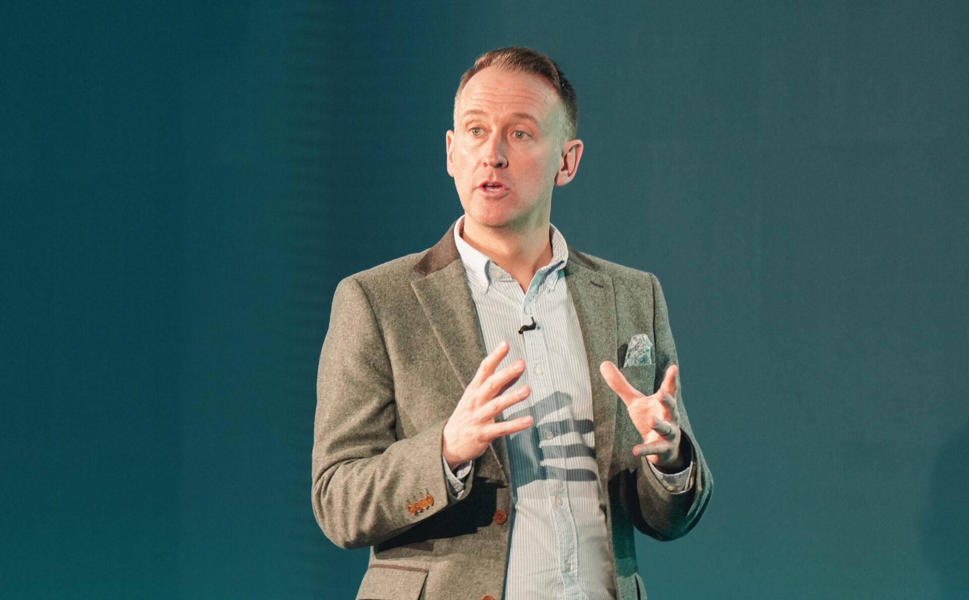 Simon Lynch speaking at a podium against a teal background, gesturing with both hands whilst presenting.
