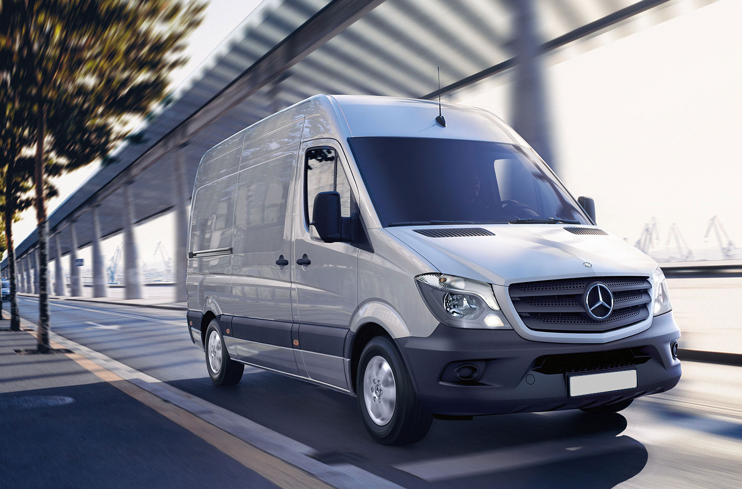 Silver Mercedes-Benz van driving on urban road beneath modern architecture and pergola structure.