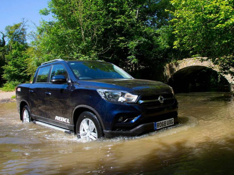 Blue SsangYong Musso pickup truck fording through river water beneath stone bridge.