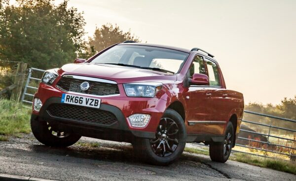 Red SsangYong Musso pickup truck parked on rural track beside farm gate, front three-quarter view.