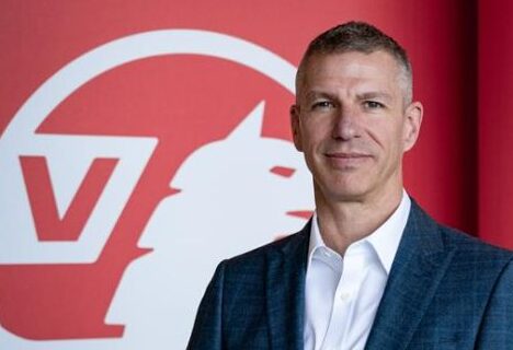 Steve Catlin in dark blazer and white shirt standing in front of red Vauxhall logo background.