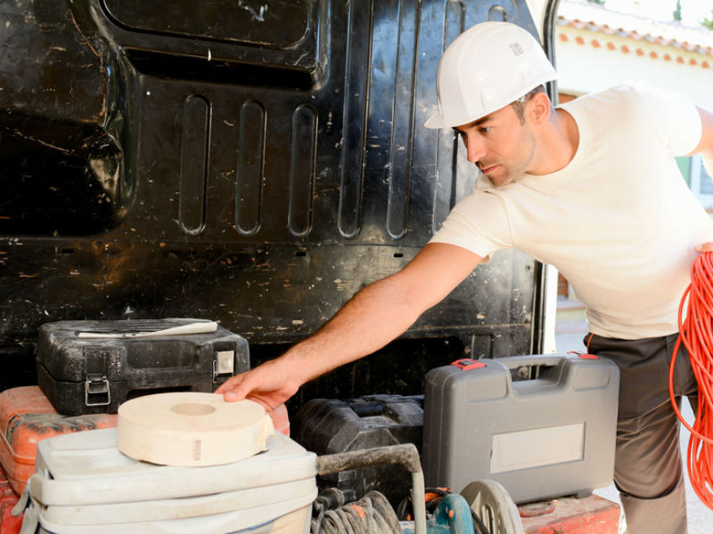 Tradesperson in white hard hat and shirt loading tools and equipment into van, organising white plastic storage boxes and…